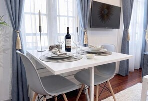 Mesa de comedor preparada con platos, vasos y una botella, rodeada de sillas grises y cortinas.
