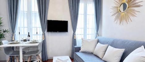 Un salon moderne avec un canapé gris, une table à manger avec des chaises, un téléviseur à écran plat fixé au mur et un miroir décoratif en forme de soleil.