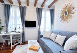 Una sala moderna con un sofá gris, una mesa de comedor con sillas, un televisor de pantalla plana montado en la pared y un espejo decorativo con diseño de sol.