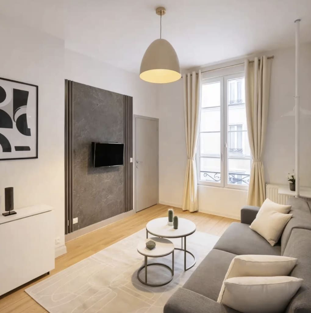 A modern living room with a sofa, a coffee table, a TV mounted on a grey wall, and a window with curtains.
