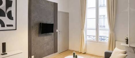 Un salon moderne avec un canapé, une table basse, un téléviseur fixé au mur gris et une fenêtre avec des rideaux.