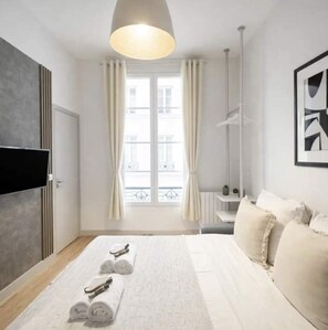Ein Hotelzimmer mit Bett, Flachbildfernseher, Fenster mit Vorhängen, Nachttisch mit Lampe und einem gerahmten Bild an der Wand.
