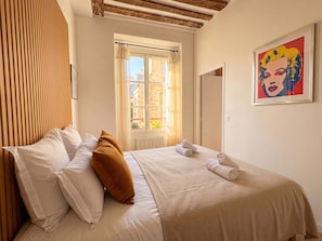 Ein Hotelzimmer mit Bett, Kissen, einem Fenster mit Vorhängen, einer Heizung, einem gerahmten Pop-Art-Gemälde und einer Tür.
