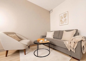 Una sala de estar con un sillón estampado, un sofá gris, una mesa de centro redonda y una obra de arte abstracta enmarcada en la pared.
