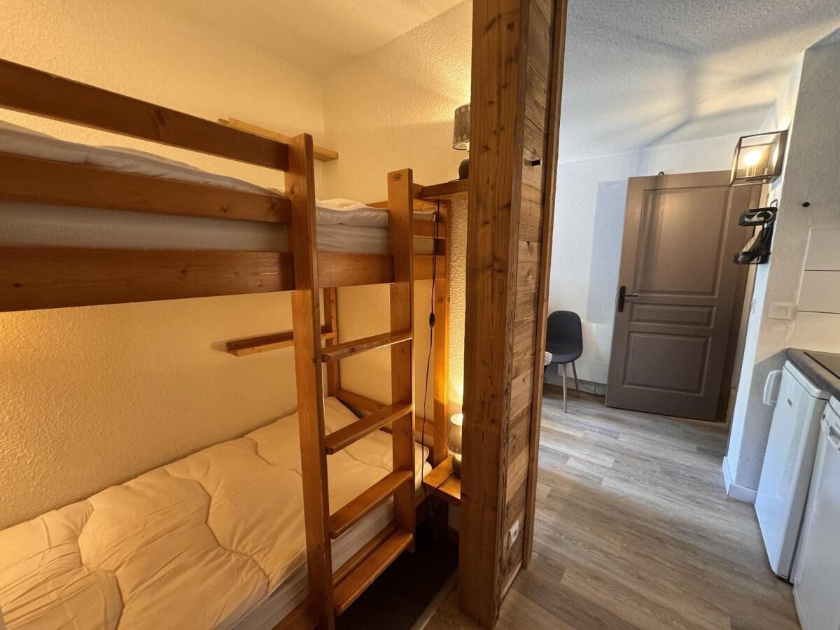 A bunk bed with a wooden frame, a small table with a lamp, a chair, and a kitchenette area with a refrigerator and a stove.