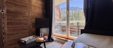 A modern hotel room with a wooden wall, a large window with a view of mountains, a bed, a small table with breakfast items, and a magazine.