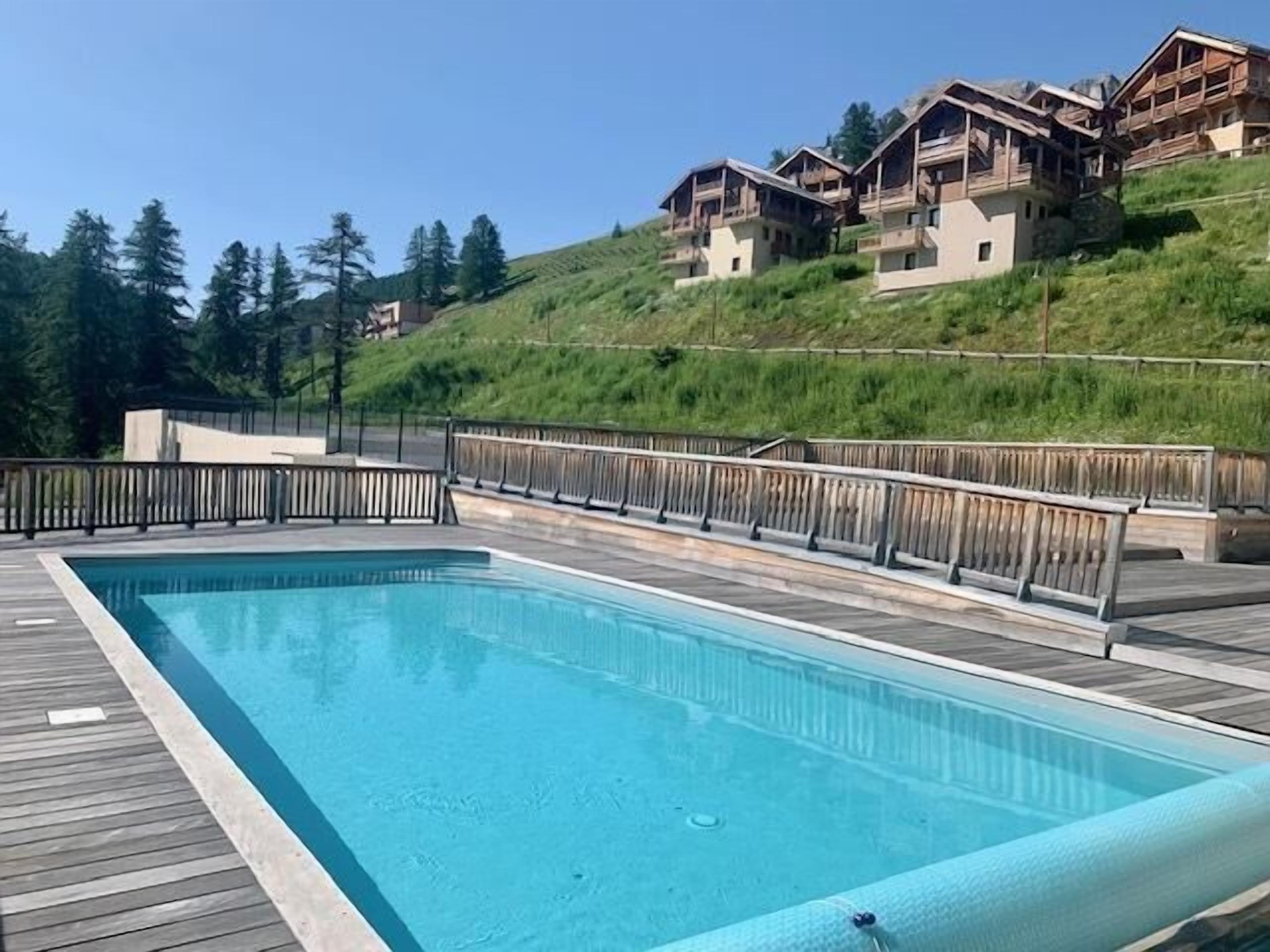 A rectangular swimming pool with a wooden deck and metal railing, surrounded by a grassy hillside and wooden buildings.
