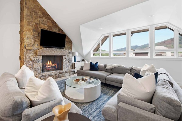 A modern living room with a stone fireplace, a large window, and a cozy seating area. - Solaris PHFW - 3 Bedroom + Den (Vail)