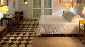 Ein Hotelzimmer mit Schachbrettboden, einem Bett mit weißen Leinen, einem Schreibtisch mit Lampe und gerahmten Bildern an den Wänden.