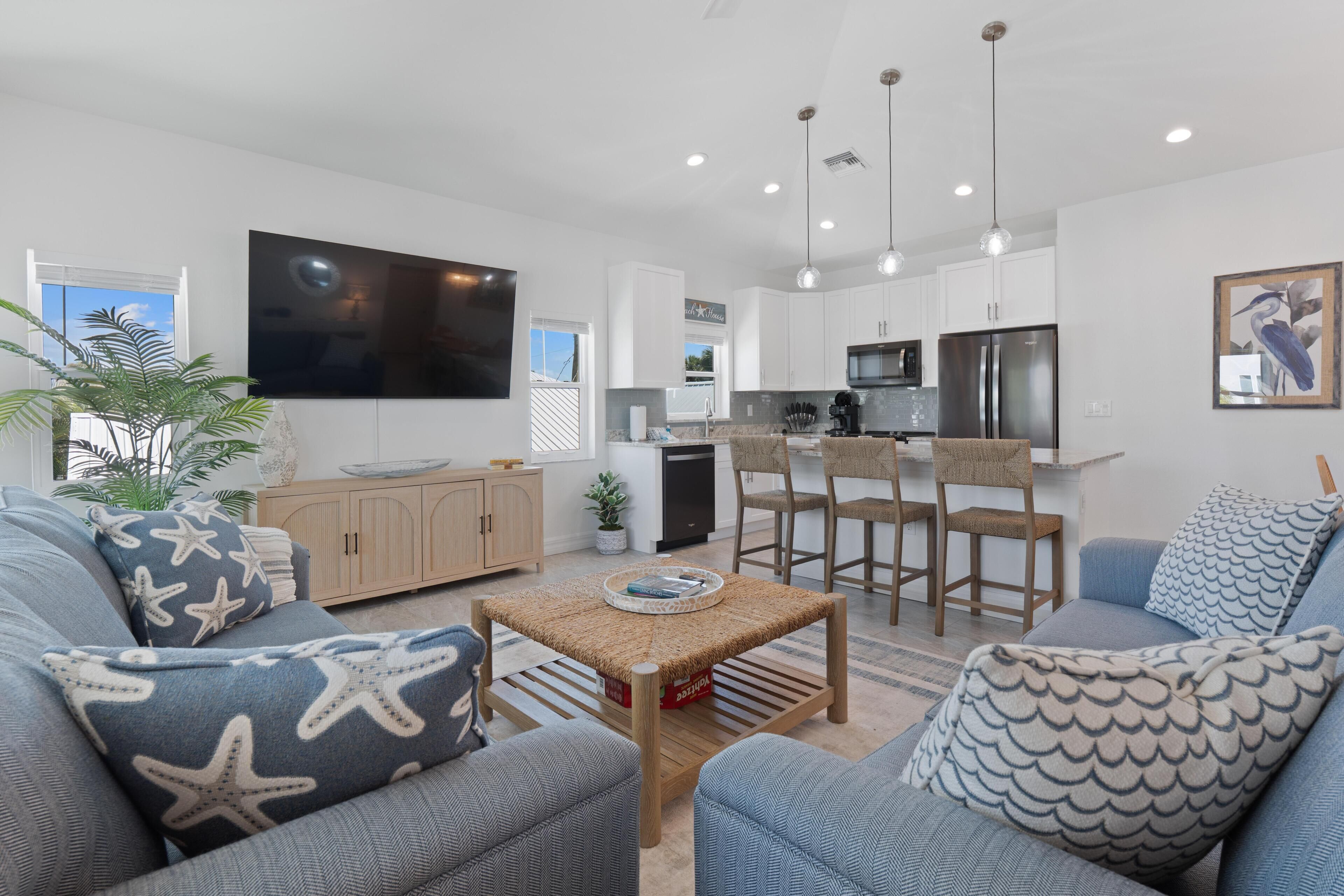 A modern living room with a large flat-screen TV, a kitchen with white cabinets, and a dining area with bar stools.