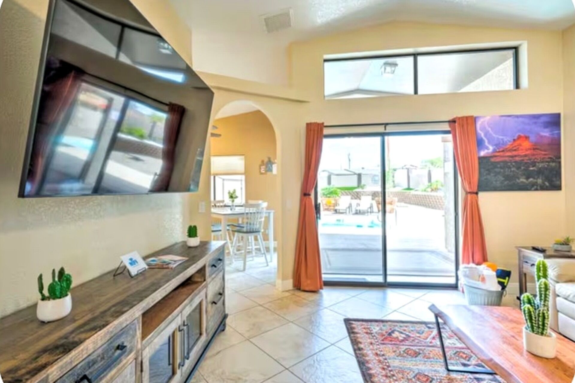 A spacious living area with a flat-screen TV mounted on the wall, a dining table with chairs, a coffee table, and a large window with curtains.