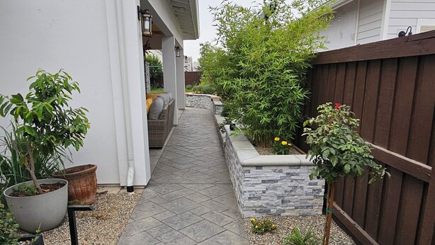 A paved pathway leading to a covered outdoor seating area with a white building on one side and a wooden fence on the other.