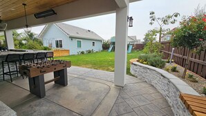 An outdoor patio area with a foosball table, seating, and a view of a backyard with a house, a slide, and a wooden fence.