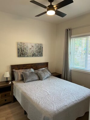 Ein Schlafzimmer mit einem Bett, einem Deckenventilator, einem Nachttisch, einer Lampe, einem Fenster mit Jalousien und einem Bild an der Wand.