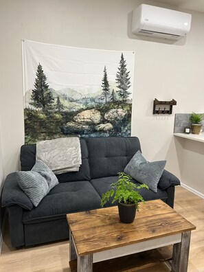 Ein Wohnzimmer mit einem dunkelgrauen Sofa, einem Couchtisch aus Holz mit einem Topf und Pflanze, einer Wandweberei mit Berglandschaft und einer wandmontierten Klimaanlage.
