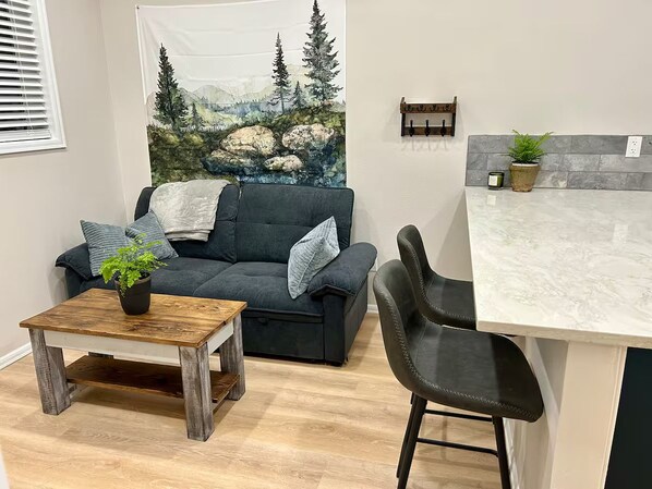 Ett mysigt vardagsrum med en mörkgrå soffa, ett träbord med en växt och en svart stol. En köksdel med en bänkskiva i marmor och en krukväxt syns.