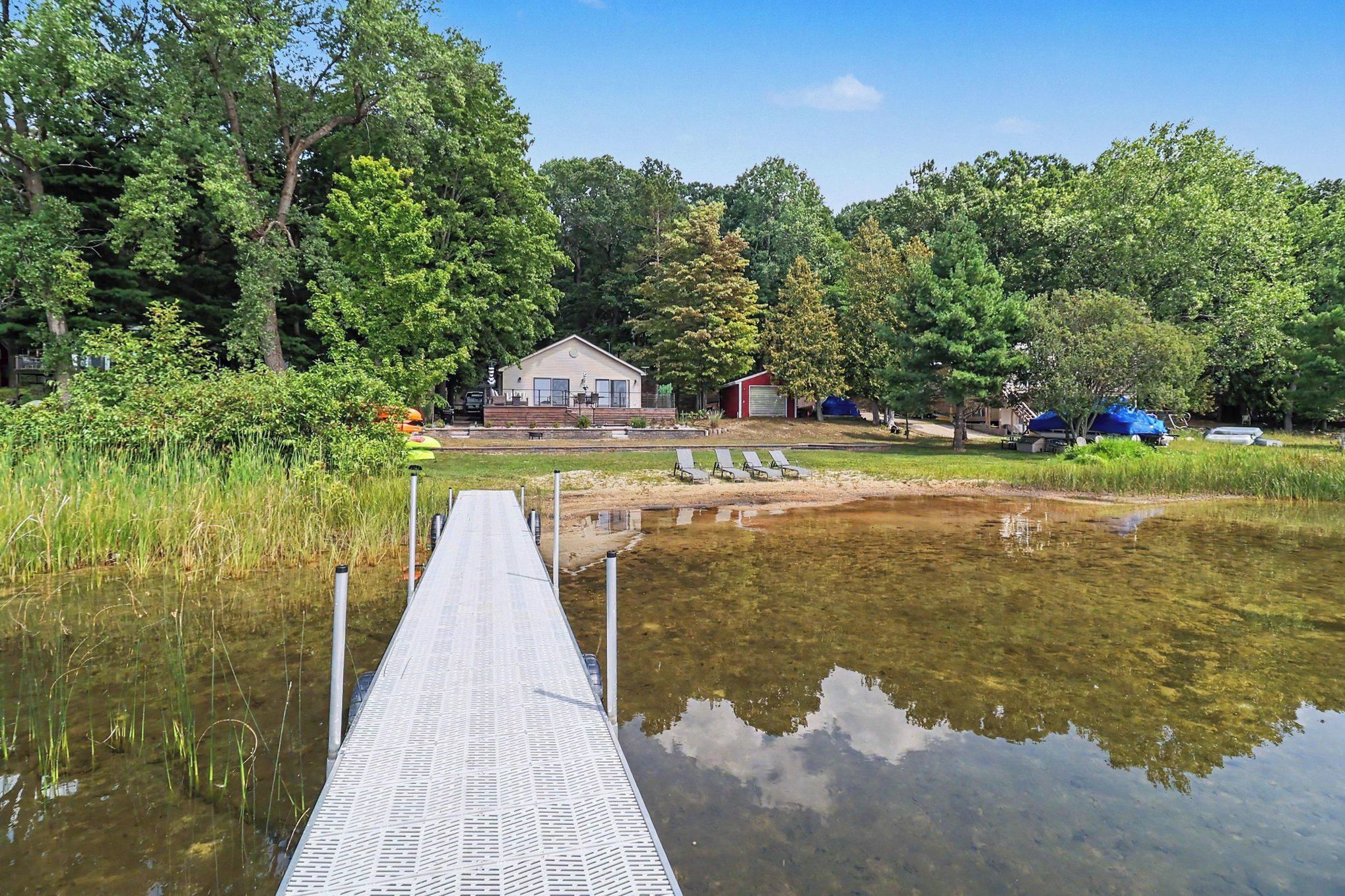 A wooden dock leading to a house with a pool, surrounded by trees and a clear sky.