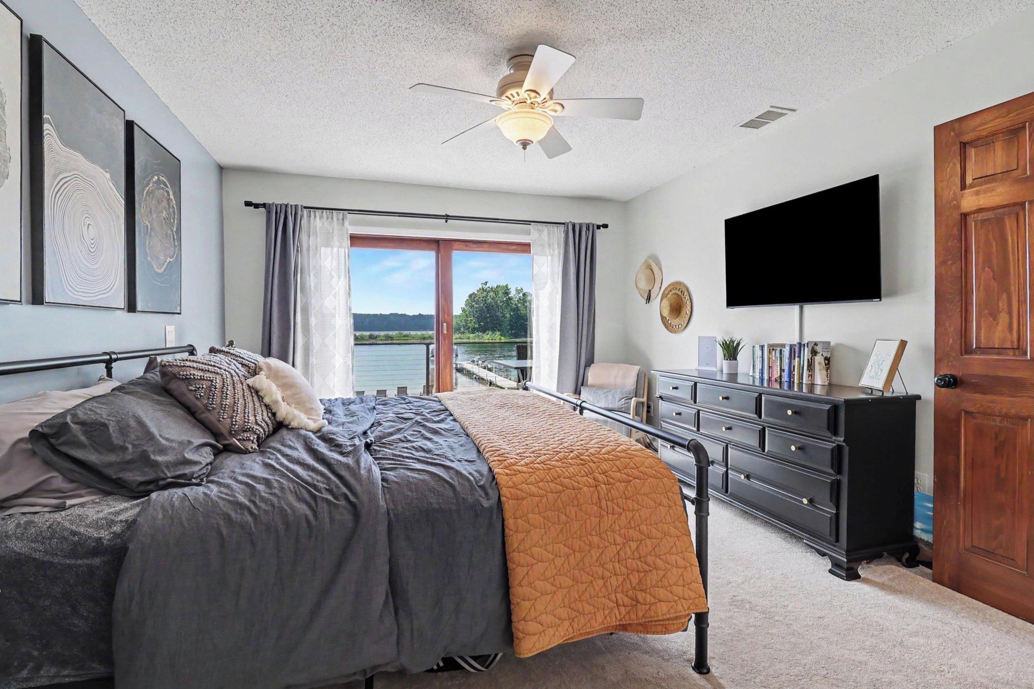 A bedroom with a bed, a dresser, a television, and a view of a body of water through a sliding door.