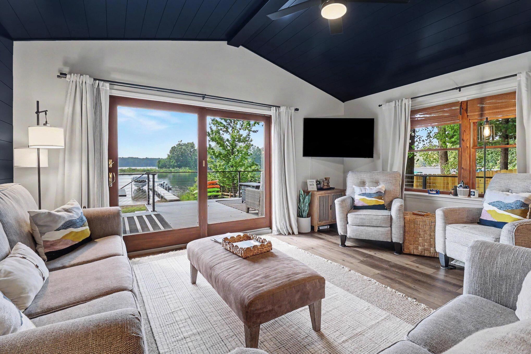 A spacious living room with a large sliding glass door leading to an outdoor deck, a comfortable sofa set, a flat-screen TV mounted on the wall, and a cozy seating area with a cushioned bench.