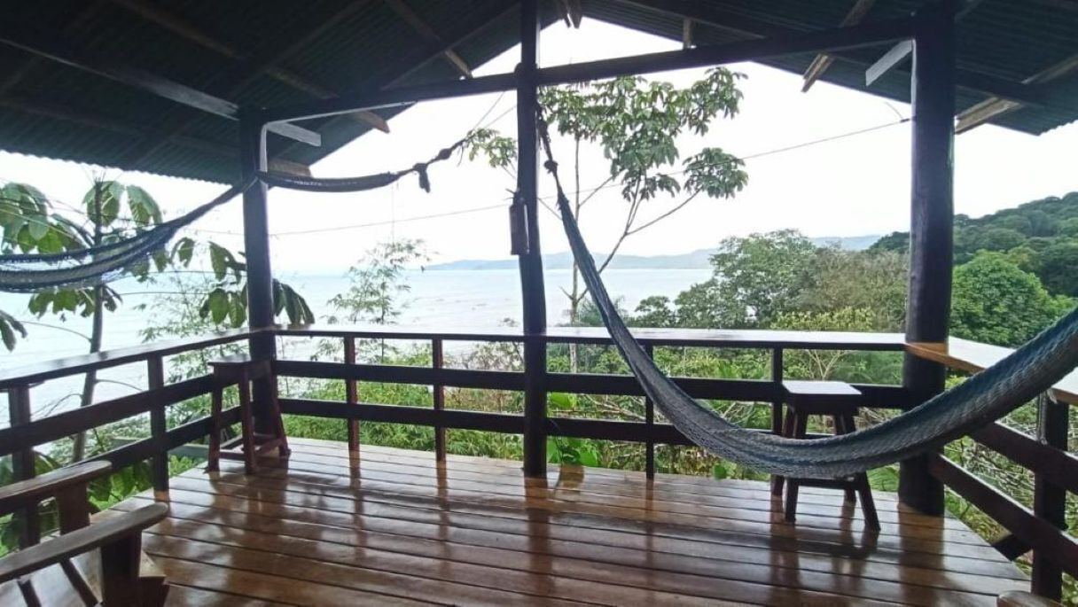Une terrasse en bois avec un hamac, des rampes en bois et une vue sur une étendue d’eau et la verdure.