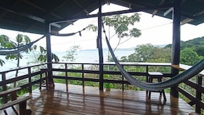 Una terraza de madera con una hamaca, barandillas de madera y vistas a un cuerpo de agua y follaje.
