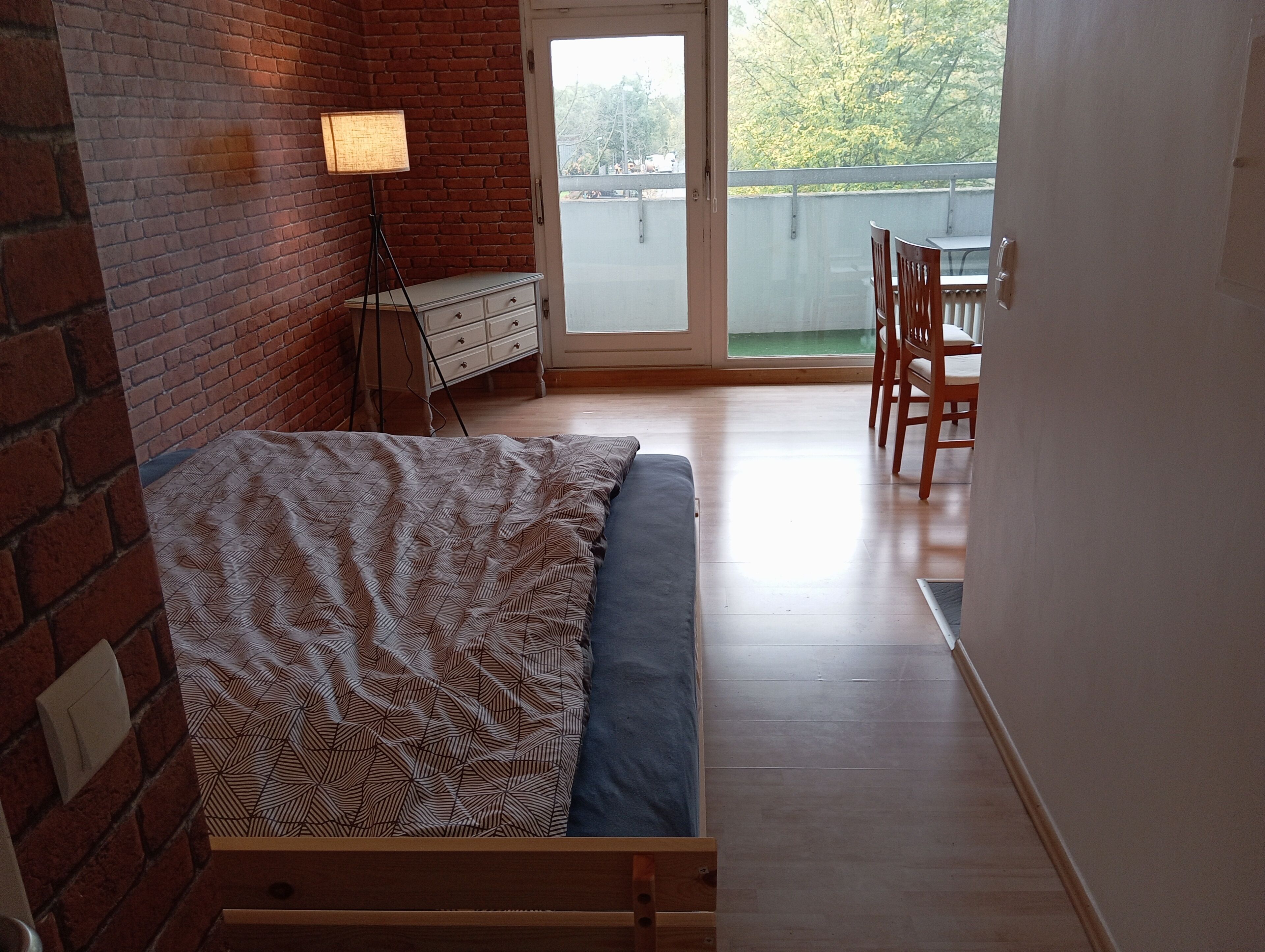 A bedroom with a bed, a dresser, a lamp, and a view of the outside.
