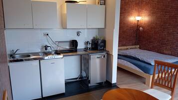A compact kitchenette with a sink, stove, and dishwasher, a bed with a brick wall backdrop, and a dining area with a wooden table and chair.