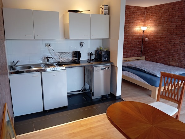 A compact kitchenette with a sink, stove, and dishwasher, a bed with a brick wall backdrop, and a dining area with a wooden table and chair.
