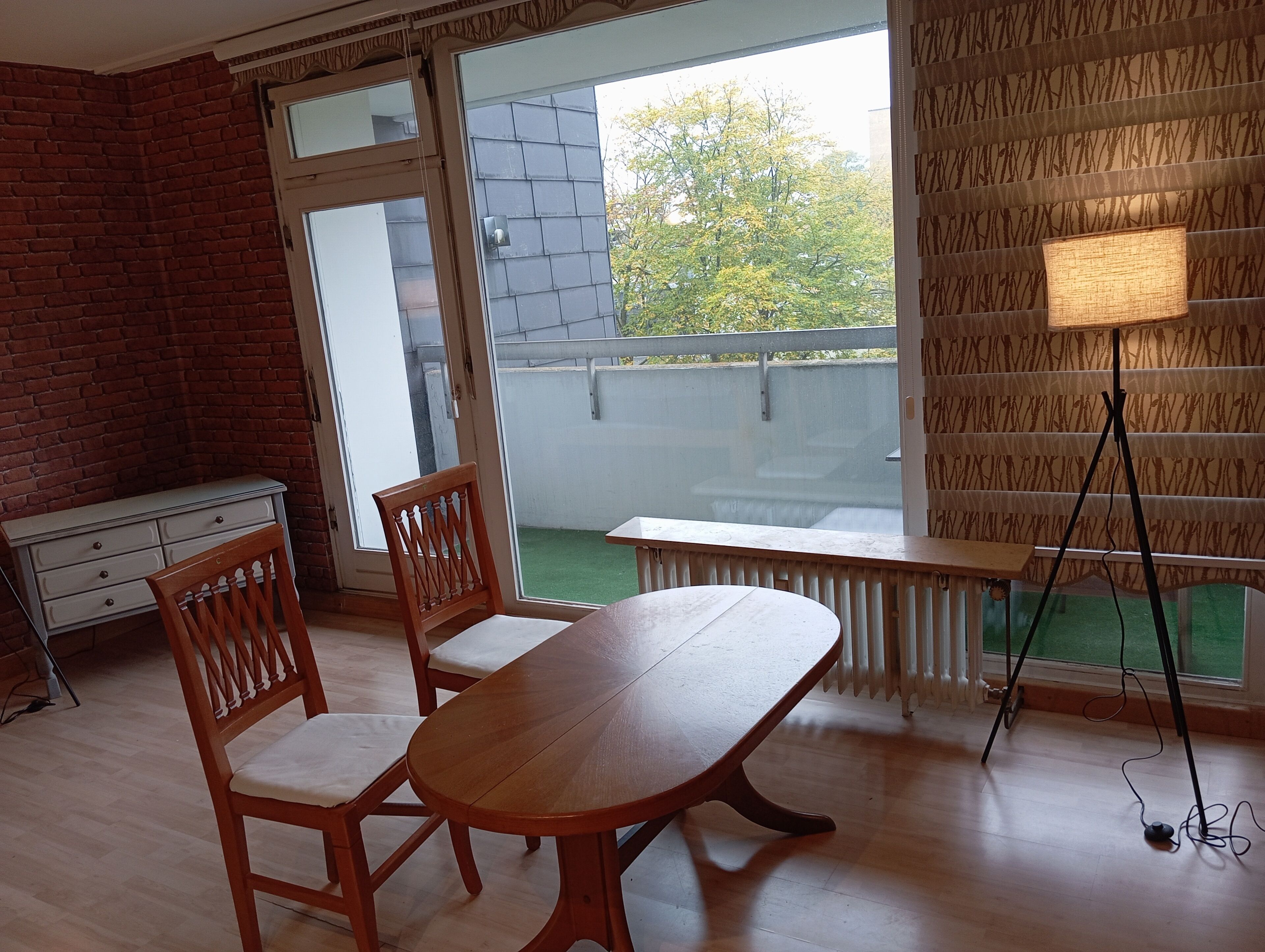 A room with a wooden table and chairs, a radiator, a bench, and a view of a balcony with a railing.
