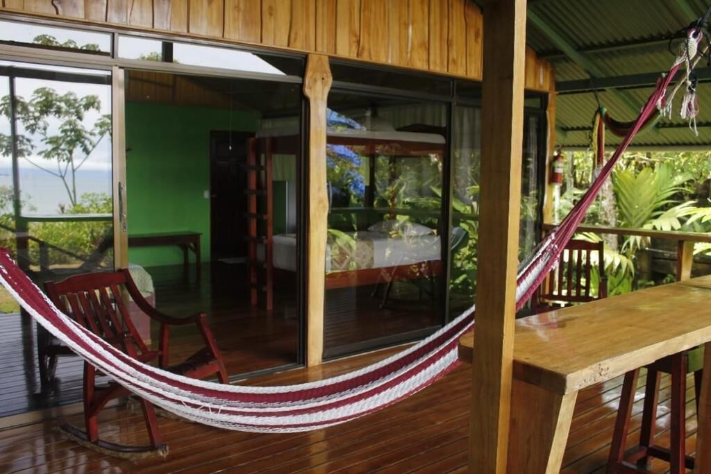 A wooden deck with a hammock, a table, and a glass door leading to a room with a bed.