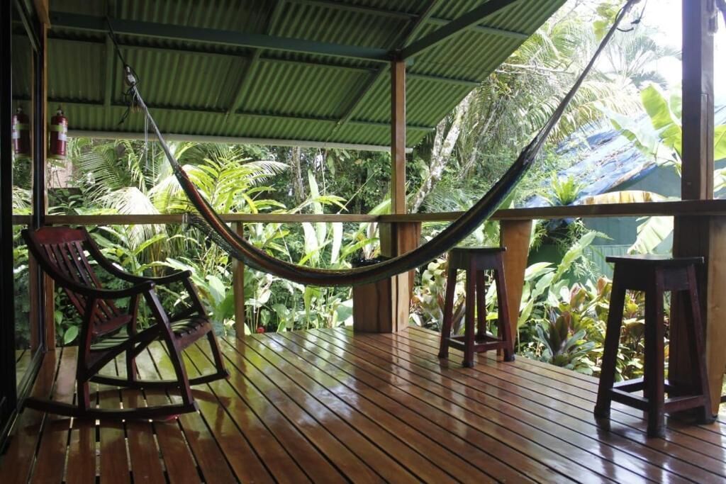 A wooden deck with a hammock, rocking chairs, and a view of lush tropical vegetation.