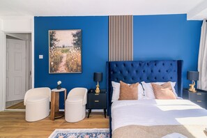 Un dormitorio con una cama grande, dos mesitas de noche, una mesilla, una silla, una pequeña mesa con un florero y un cuadro en la pared.