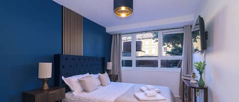A bedroom with a large bed, a bedside table, a lamp, a window with curtains, and a wall-mounted TV.