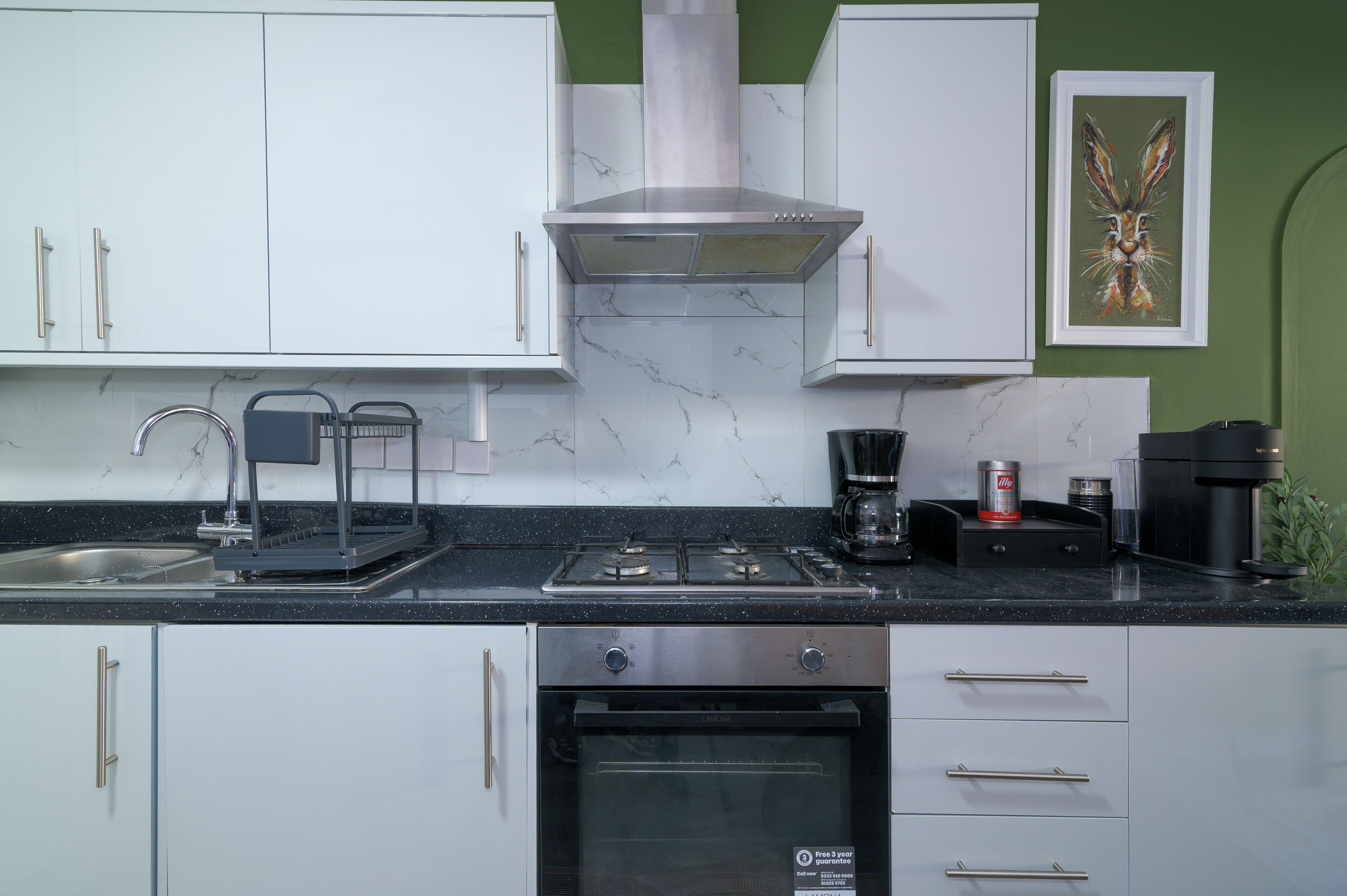 Une cuisine moderne avec des armoires blanches, un comptoir noir, une cuisinière en acier inoxydable, une machine à café et une œuvre d’art encadrée accrochée au mur.