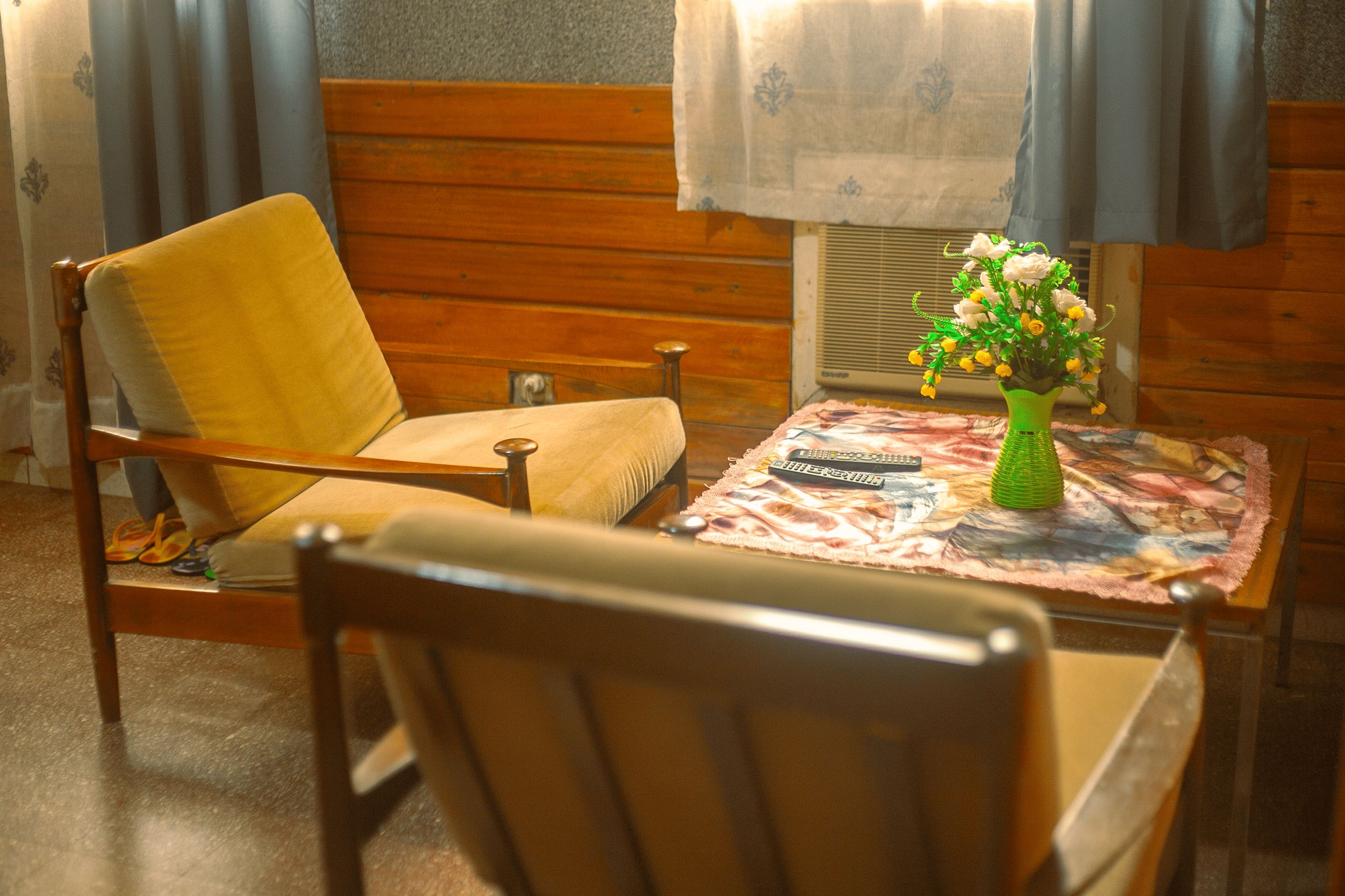 Una habitación con una silla de madera, una mesa con mantel floreado, un jarrón verde con flores y un aire acondicionado de pared.