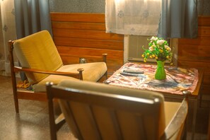 A room with a wooden chair, a table with a floral tablecloth, a green vase with flowers, and a wall-mounted air conditioner.