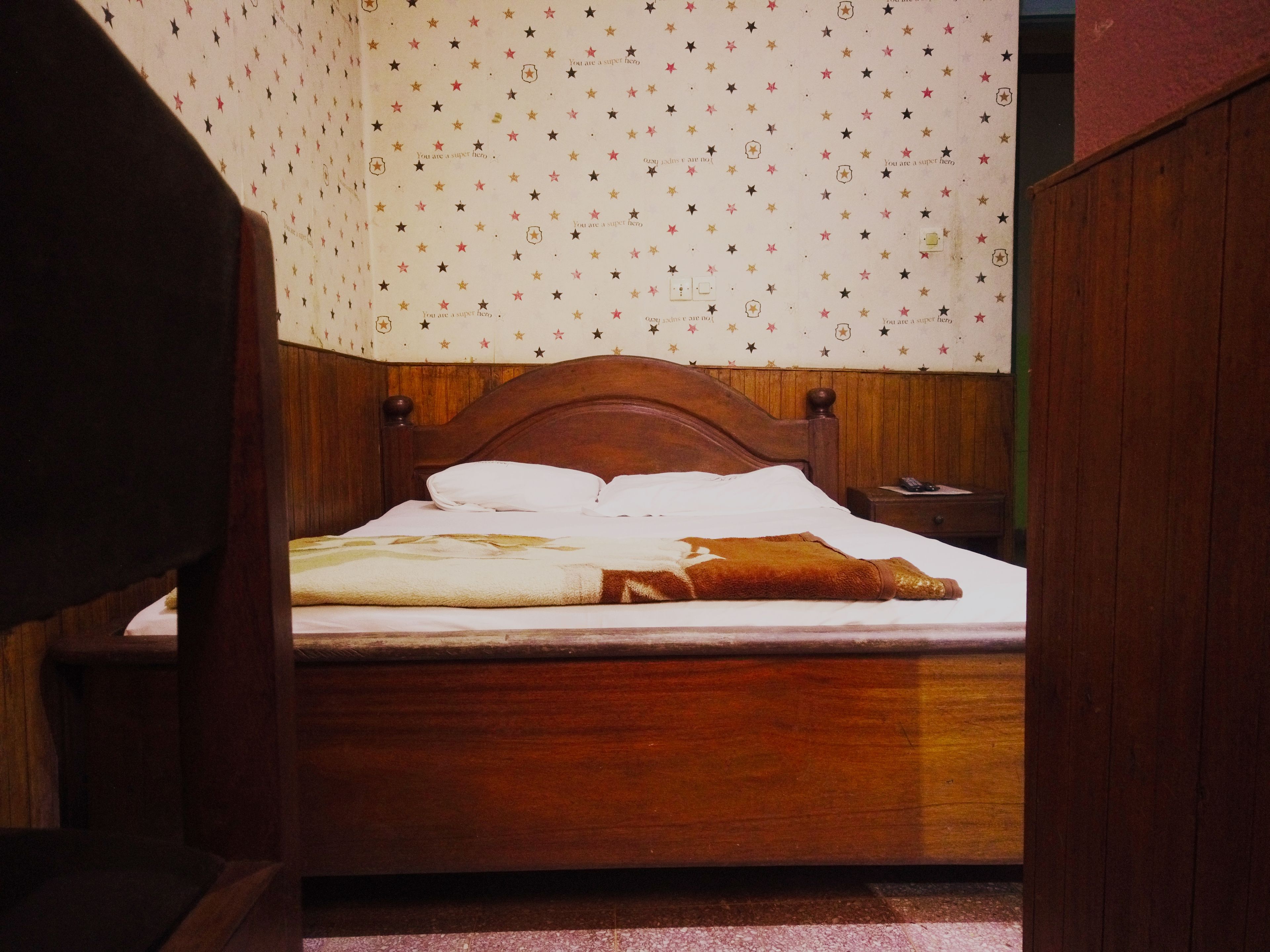Una cama de madera con colchón blanco y manta marrón, cabecera de madera y pared con diseño de estrellas.