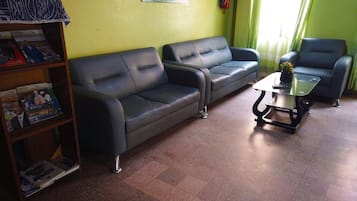 Una habitación con dos sofás negros, una mesa de centro de cristal, una estantería con revistas, un extintor y una ventana con cortinas.