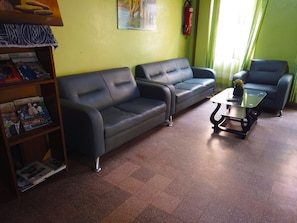 A room with two black sofas, a glass coffee table, a bookshelf with magazines, a fire extinguisher, and a window with curtains. - Hôtel Belle Vue Yaounde (Yaoundé)