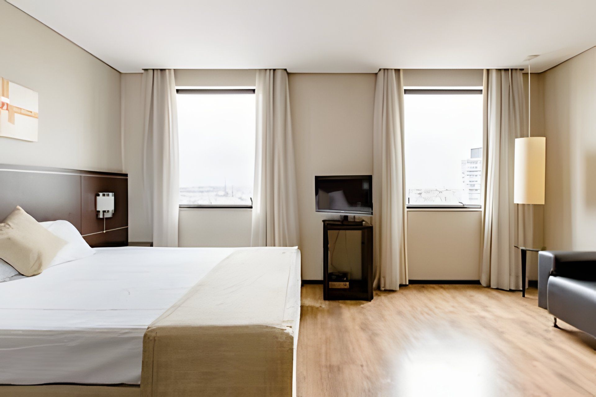 A hotel room with a large bed, a TV on a stand, a desk with a lamp, and a view of the city through the windows.