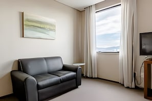 Una habitación de hotel moderna con un sofá de cuero negro, una pequeña mesa redonda, un televisor de pantalla plana y un ventanal con cortinas.