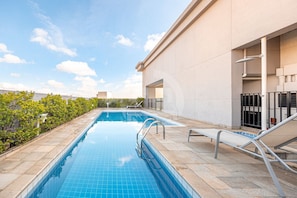 Una piscina en la azotea con cielo azul despejado, una terraza con baldosas y una barandilla de metal.