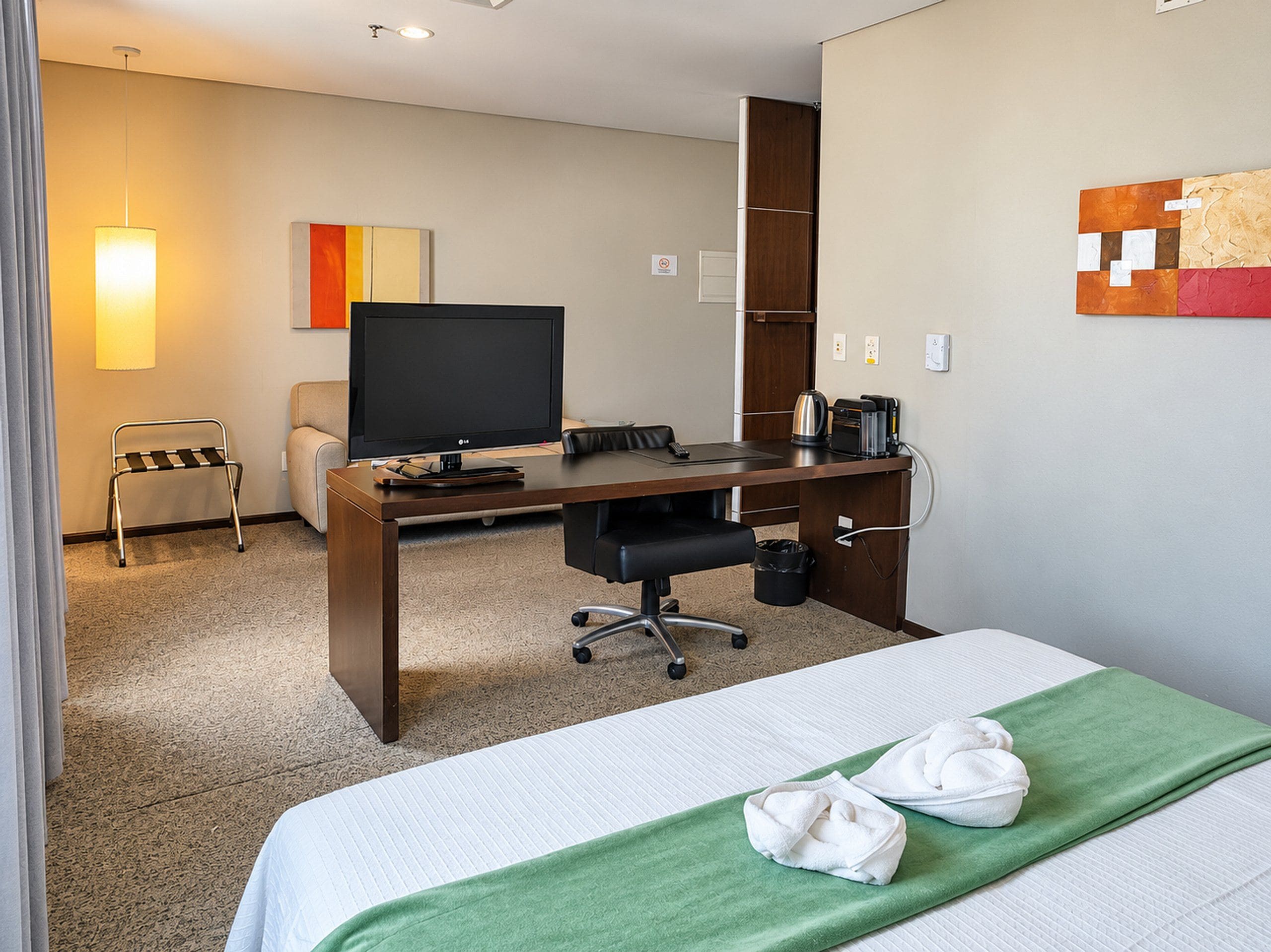 A hotel room with a bed, a desk with a computer, a chair, a lamp, and a wall art piece.