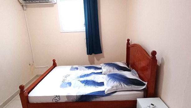 Ein Schlafzimmer mit einem hölzernen Bett, einem Nachttisch, einer weißen Schachtel, einem Fenster mit Vorhang und einer Klimaanlage.