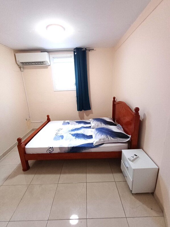 Een slaapkamer met een houten bed, een nachtkastje, een witte doos, een raam met gordijn en een airconditioning.