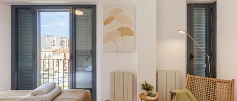 A bedroom with a bed, a chair, a radiator, a window, and a view of buildings outside.