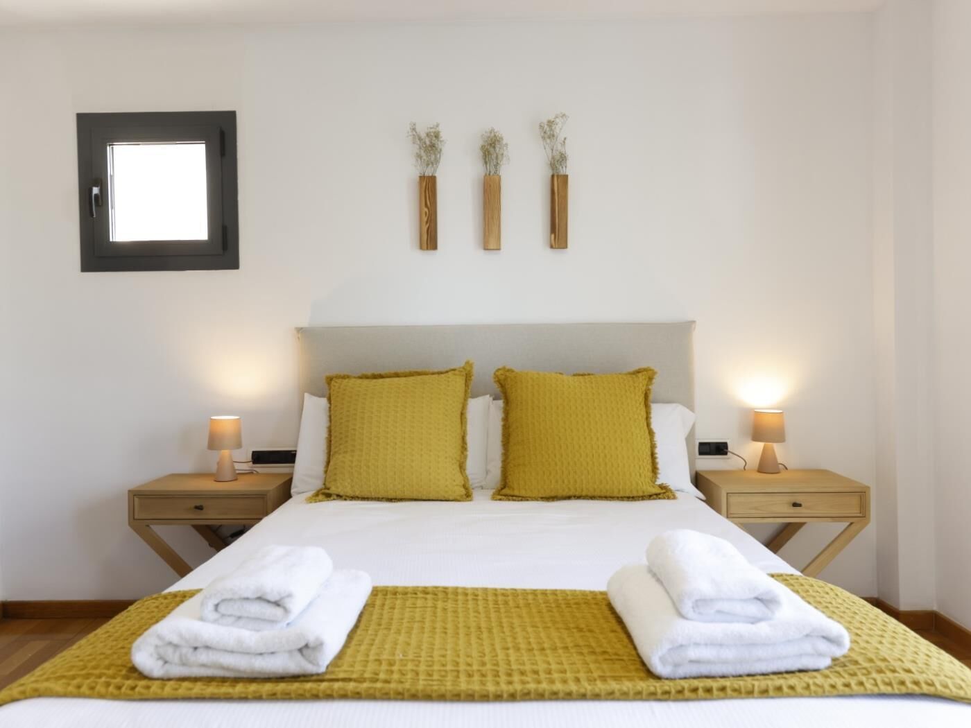 A hotel room with a bed, two bedside tables, a nightstand, a framed mirror, and three wall-mounted vases with dried flowers.