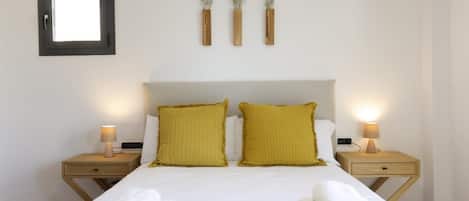 Habitación de hotel con cama, dos mesitas de noche, una lámpara de noche, un espejo en marco y tres jarrones de pared con flores secas.