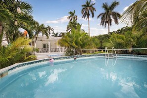A swimming pool with a clear blue water surface, surrounded by palm trees and a covered seating area. - Vrbo Property (Hollywood)