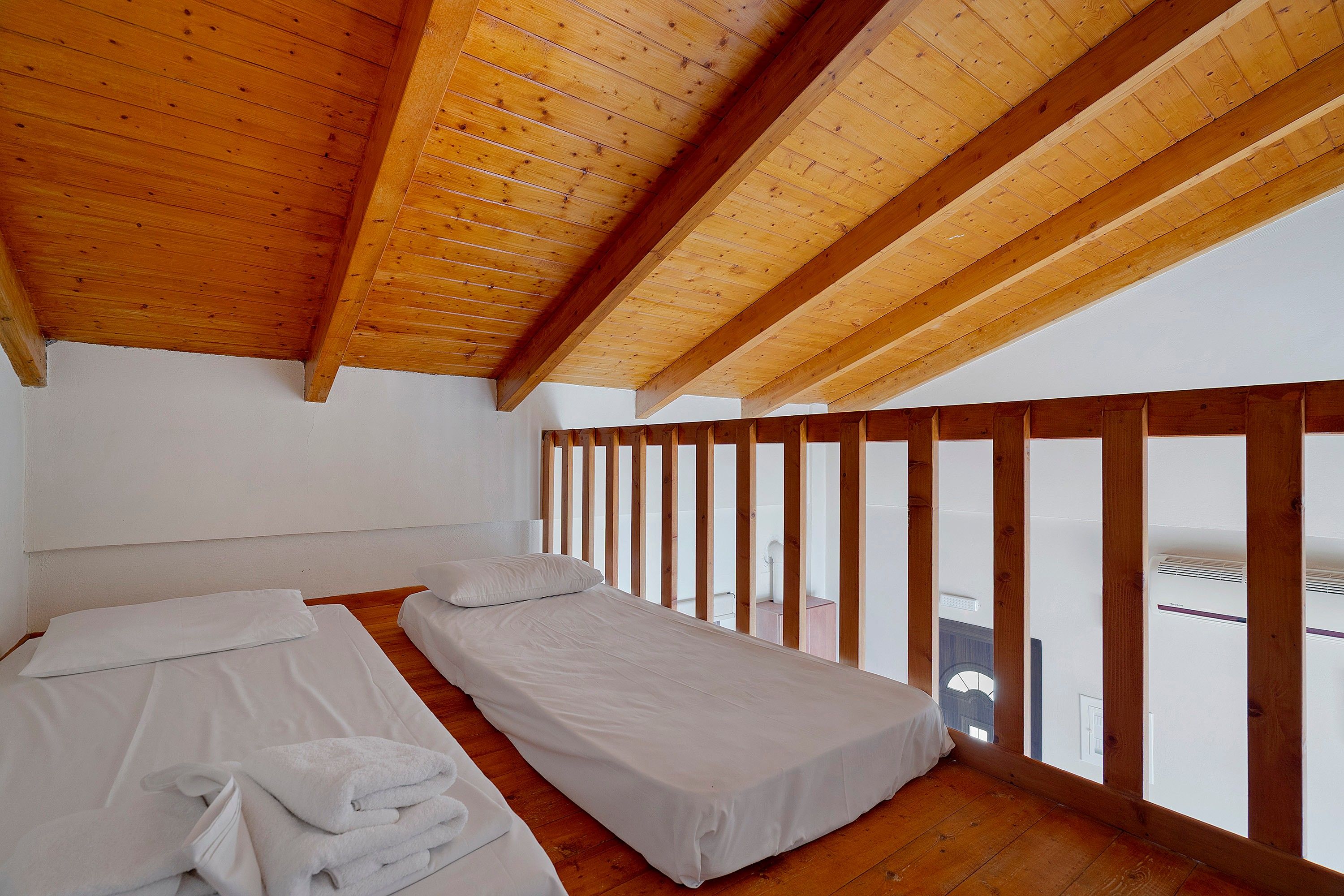 A room with two single beds, wooden beams on the ceiling, and a balcony with a wooden railing.